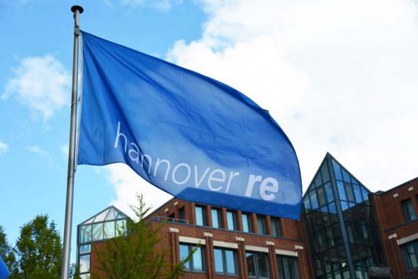 Blaue Flagge mit Aufschrift "hannover re" weht vor einem modernen Gebäude unter blauem Himmel.