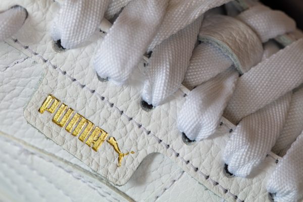 Nahaufnahme eines weißen Sneakers mit goldfarbenem Logo und weißen Schnürsenkeln.