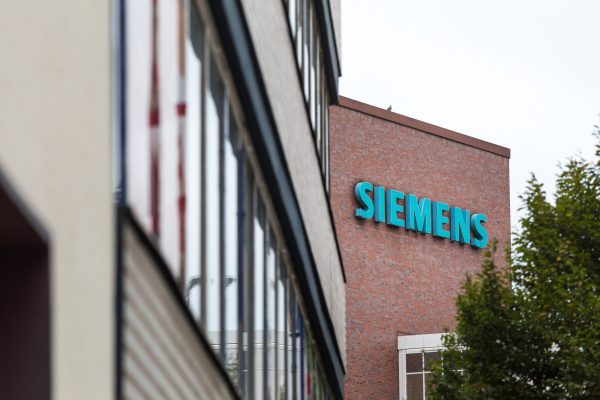 Rotes Gebäude mit blauem Schriftzug "Siemens", daneben modernes Gebäude und Baum.